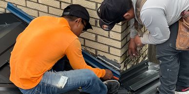 Two workers installing metal roofing near a brick wall.