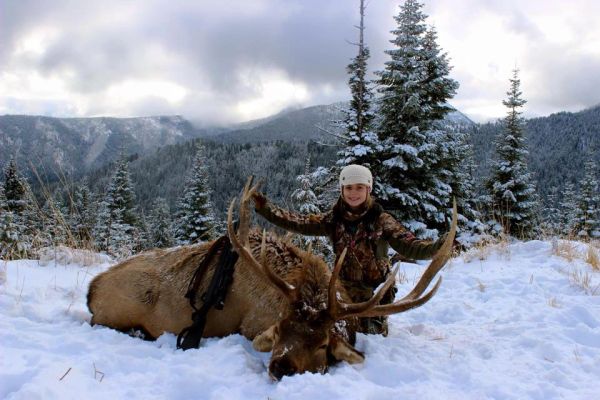 A hunter posing with a large elk in snowy mountains.