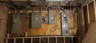 Electrical panels and wiring mounted on a plywood wall in a basement or utility room.