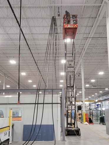 Worker on scissor lift installing cables in a large industrial warehouse.