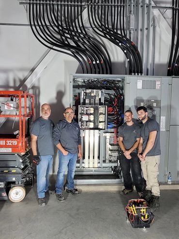 Four electricians posing in front of a complex electrical panel with organized cables.