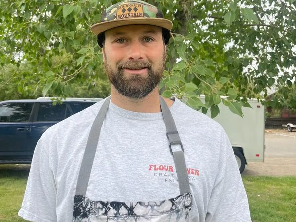 Man with beard wearing a camouflage cap and apron outdoors.