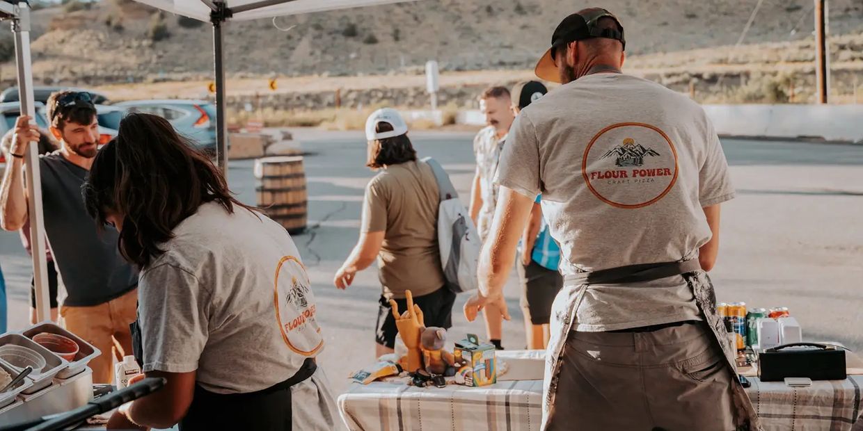 Two people wearing Flour Power Craft Pizza shirts serve food at an outdoor event.