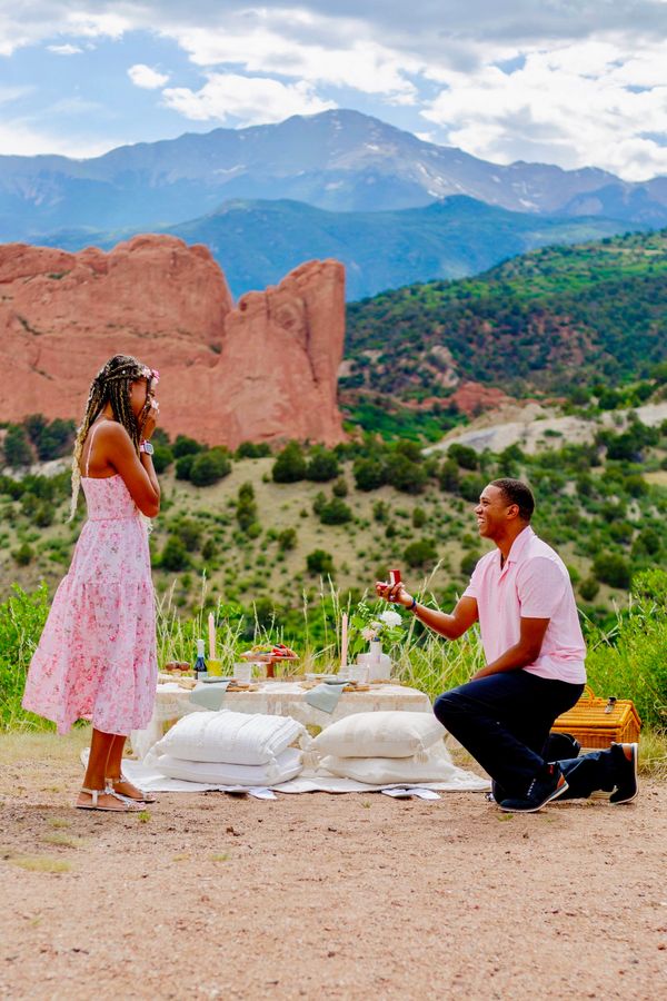 Man proposing to woman at a scenic outdoor picnic with mountains in the background.