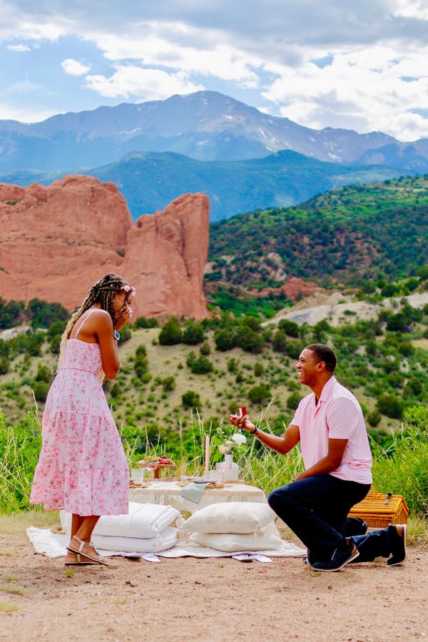Man proposes to woman during a picnic with scenic mountain backdrop.