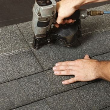 Hands using a pneumatic nail gun to install asphalt shingles on a roof.