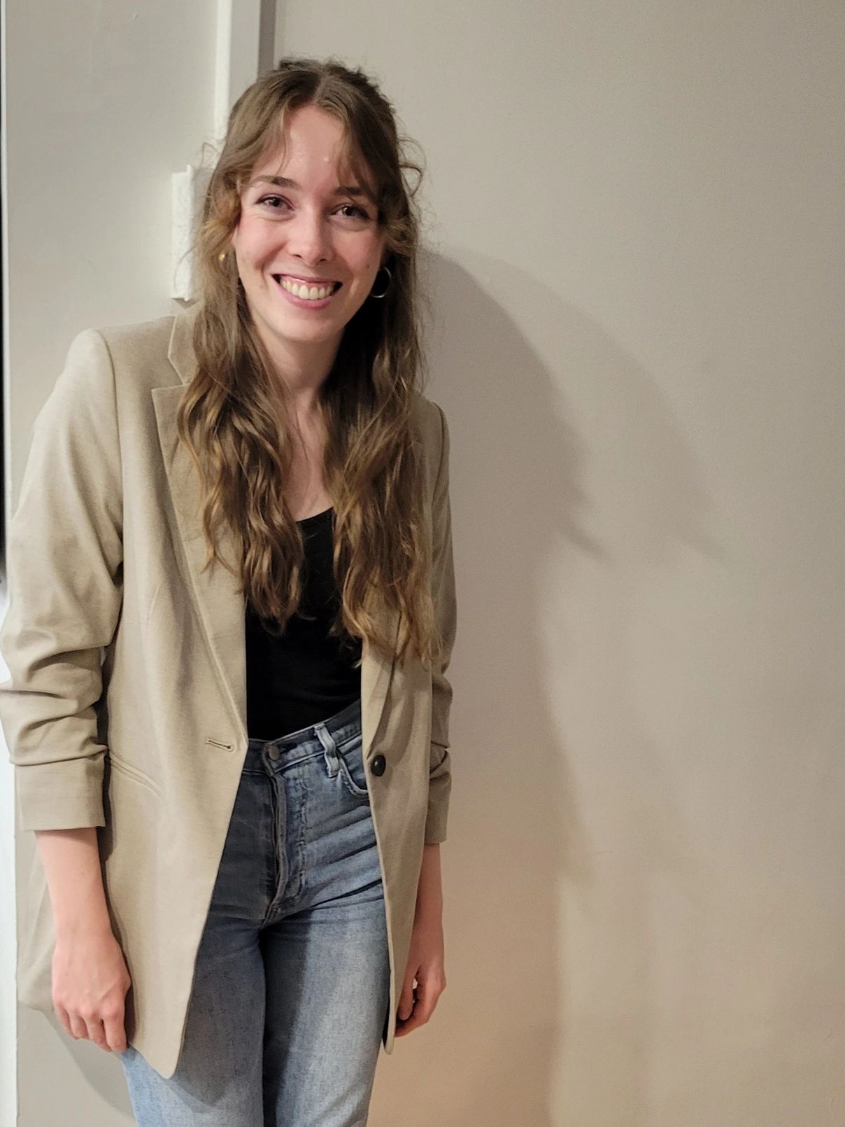 Image of Samantha McNeil leaning against a cream background. She is wearing jeans and a blazer.