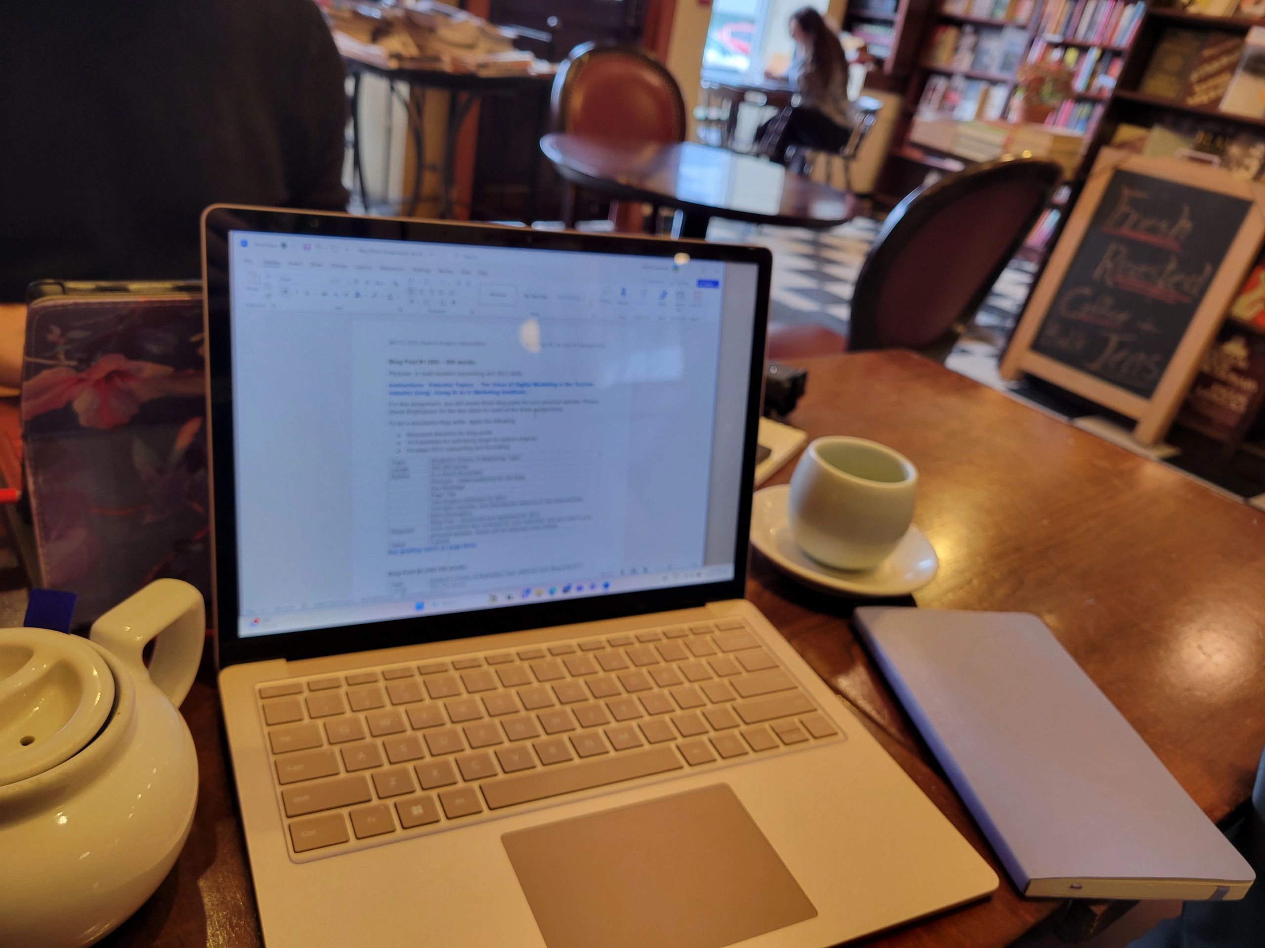 A computer rests on a table in a coffee shop. Next to the computer is a notebook and a tea cup.