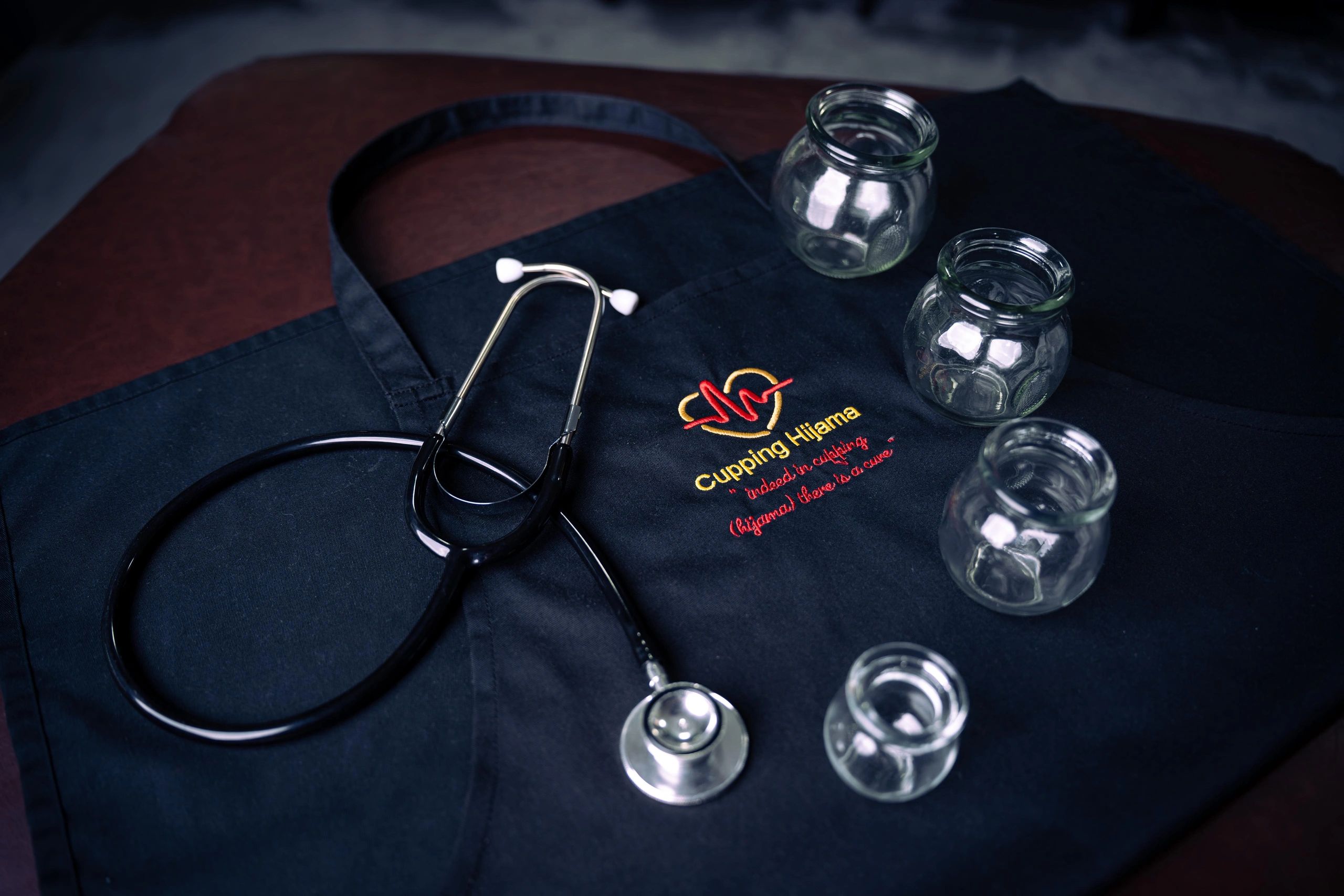 Stethoscope and cupping jars on a black apron embroidered with 'Cupping Hijama'.