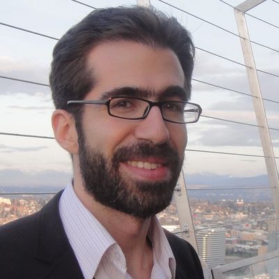 Smiling man with glasses and beard in a suit on a city balcony.