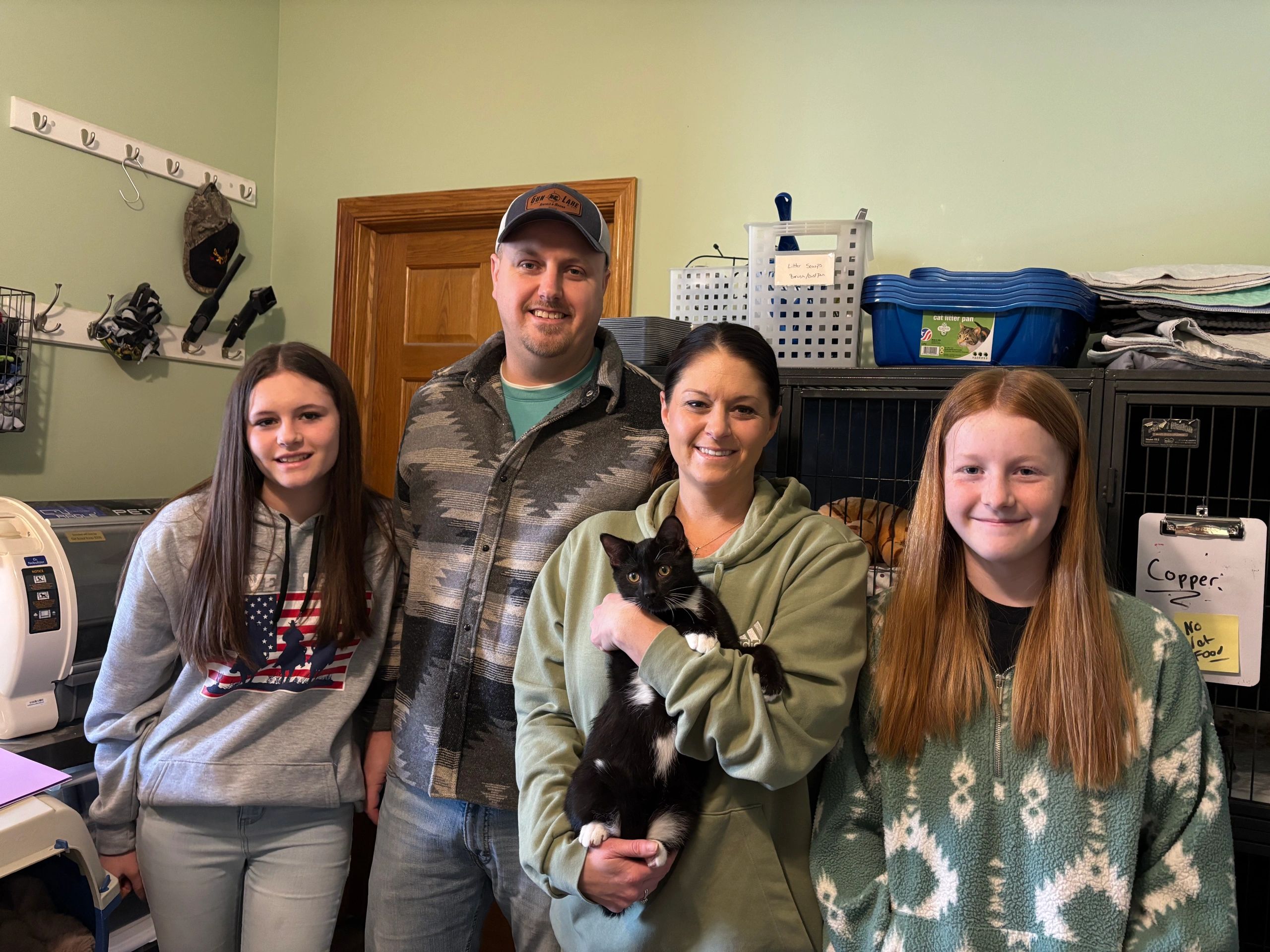 A family of four smiling indoors, with the mother holding a black and white cat.