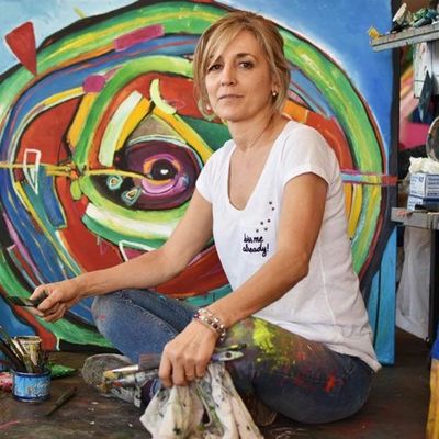 Mujer artista pintando frente a una obra colorida.