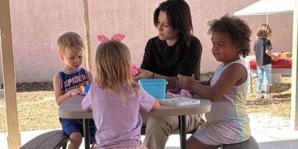 Adult and children crafting at a picnic table outdoors.