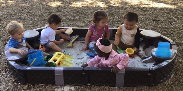 Five toddlers playing in a sandbox with toys.