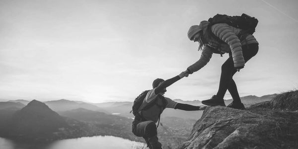 Two hikers helping each other climb a steep rock with a scenic mountain view.