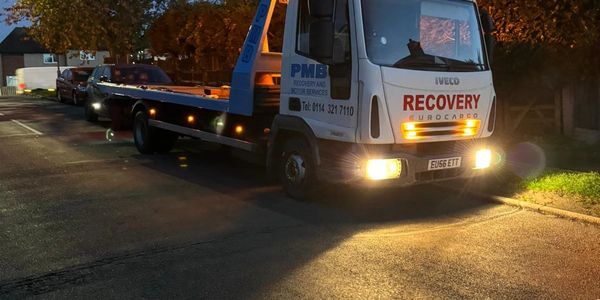 A recovery truck with headlights on parked on a street at dusk.