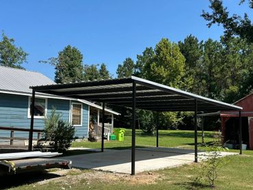 Metal carport structure on a concrete slab beside a blue house under a clear sky.