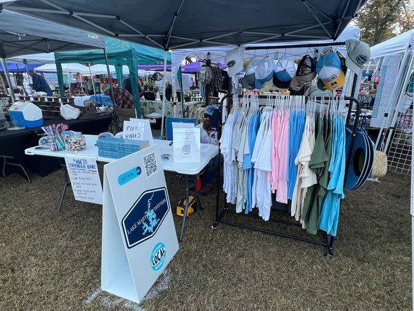 Outdoor market booth selling clothing, hats, and free Bibles with a scavenger hunt sign.