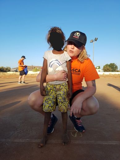 Volunteer in orange shirt kneels beside a child on a sunny outdoor court.
