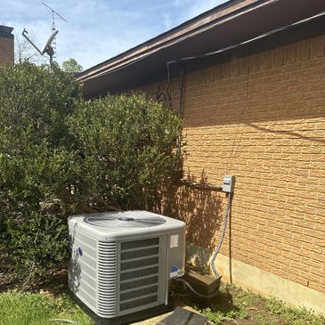 Outdoor air conditioning unit next to a brick house wall with bushes.