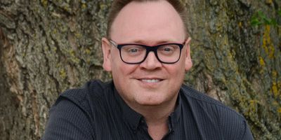 Man in glasses sitting against a tree, smiling warmly.