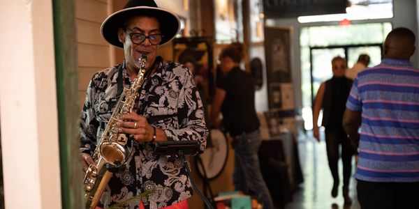 A stylish man plays saxophone indoors while people walk in the background.