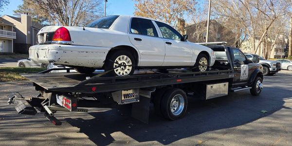 A white car with rear damage is being towed on a flatbed truck.