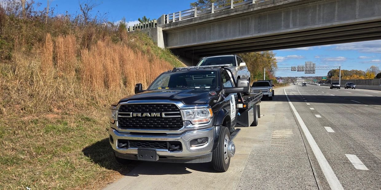 A tow truck carrying a silver SUV is parked by the highway under a bridge.
