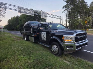 A black RAM tow truck carrying a sedan on a roadside grassy area.