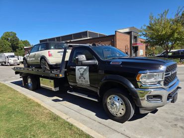 A black tow truck carrying a beige Mini Cooper on a sunny day.