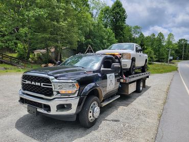 A black Ram tow truck carrying a white pickup on its flatbed on a gravel roadside.