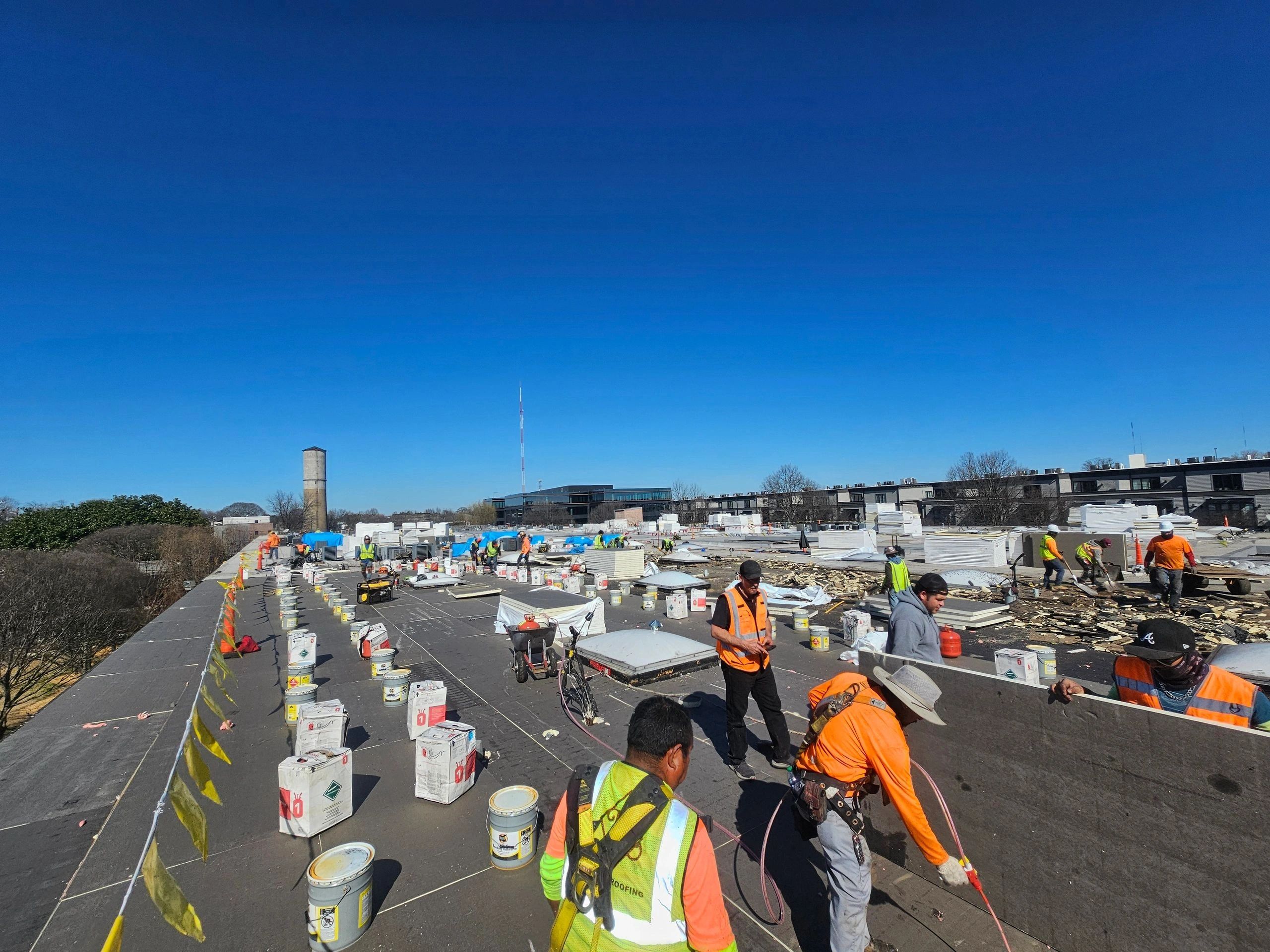 Workers in safety vests and helmets repairing a large flat rooftop under clear blue skies.