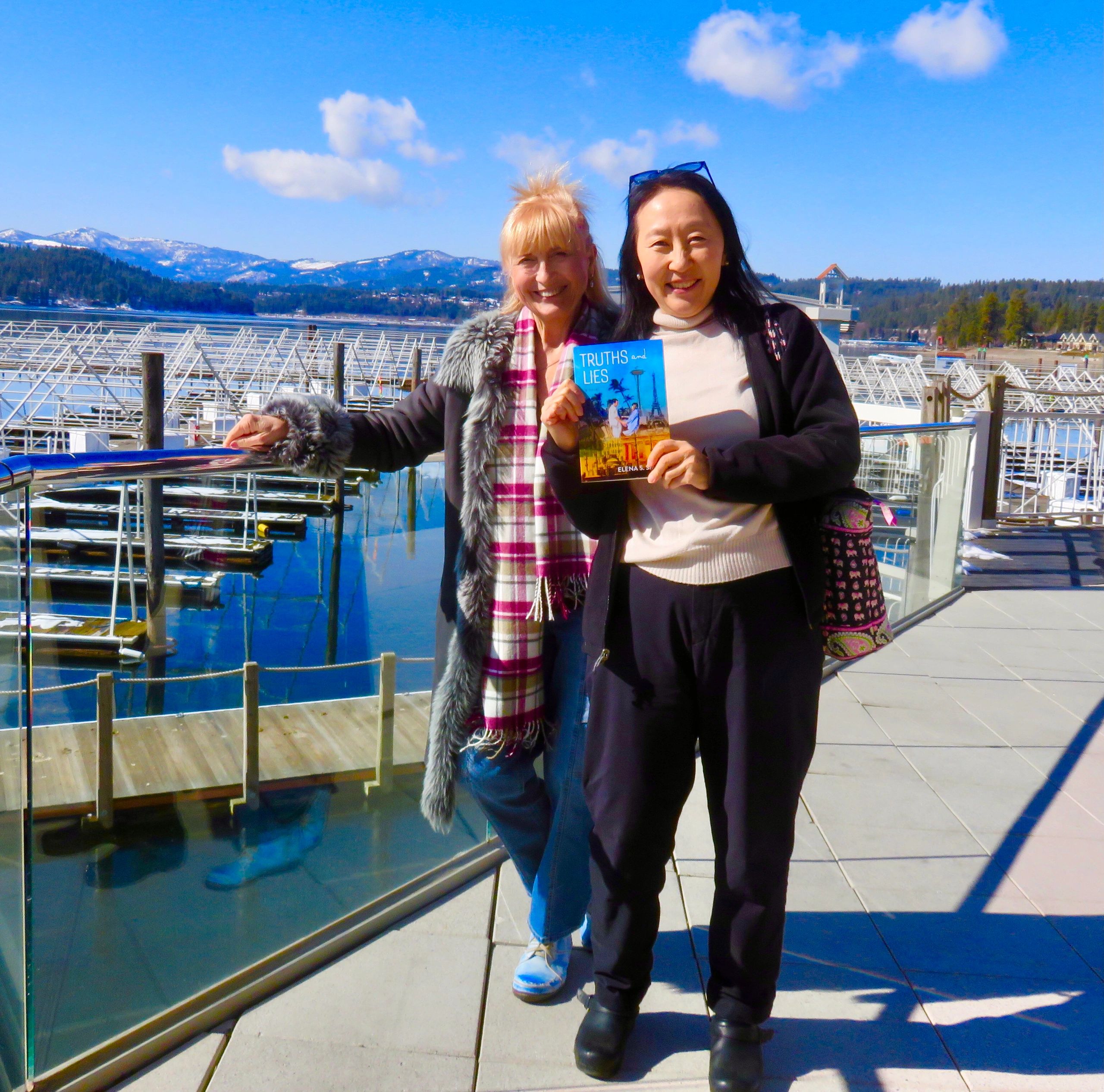Two women smiling by a marina, one holding a book titled 'Truths and Lies.'