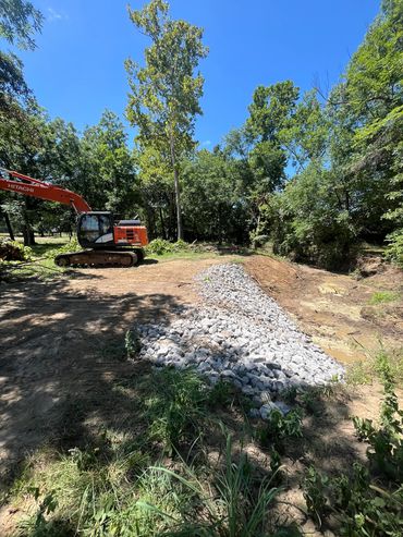 Excavator near a rocky drainage area in a wooded clearing under clear blue sky.