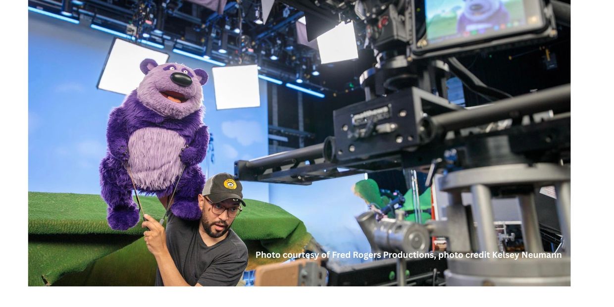 A puppeteer operates a large purple bear puppet on a TV set with cameras and lights.