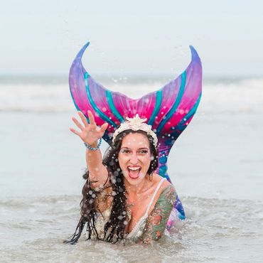 A joyful woman with a colorful mermaid tail splashes in the water at the beach.
