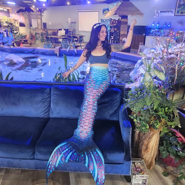 A woman dressed as a mermaid sits on a blue couch in a colorful indoor setting.