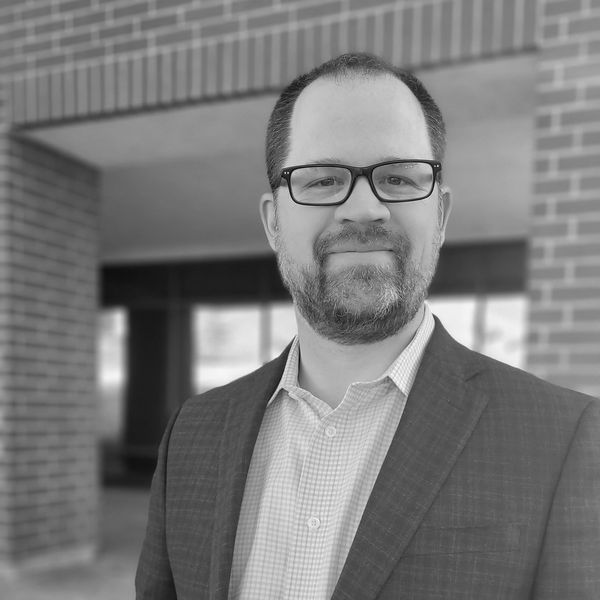 Man with glasses and beard in a suit jacket smiling outdoors.