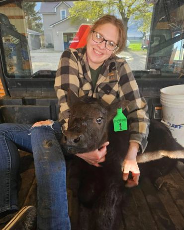 Woman wearing glasses cuddling a black calf with a green ear tag.