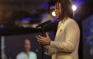 Man with braided hair sings into a microphone on stage.