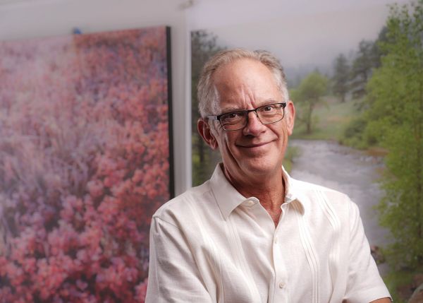 Smiling elderly man with glasses indoors, colorful art in background.