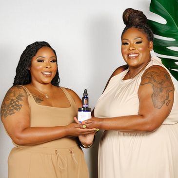 Two smiling women holding skincare products, framed by large green leaves.