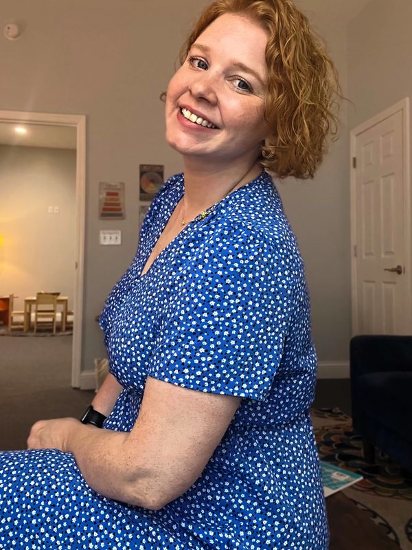 Smiling woman in a blue floral dress sitting indoors.