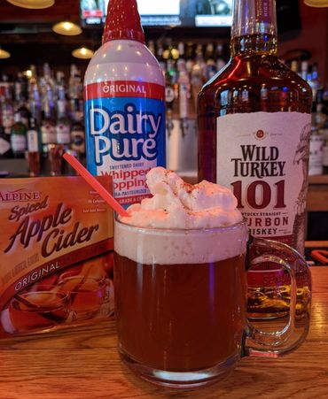 a mug of apple cider on a bartop next to a packet of apple cider and bottle of wild turkey bourbon 