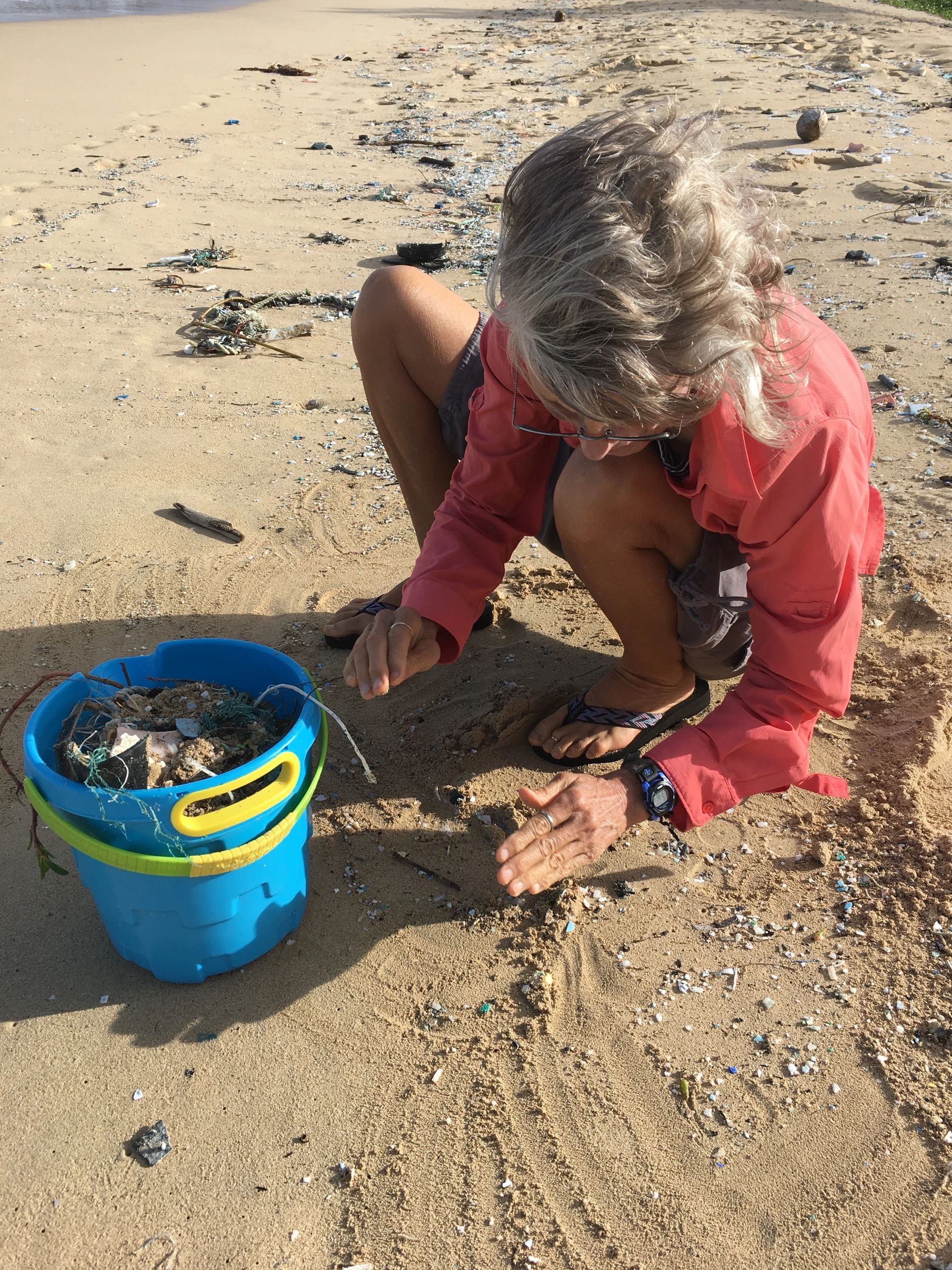Person cleaning plastic debris from a sandy beach with a blue bucket.
