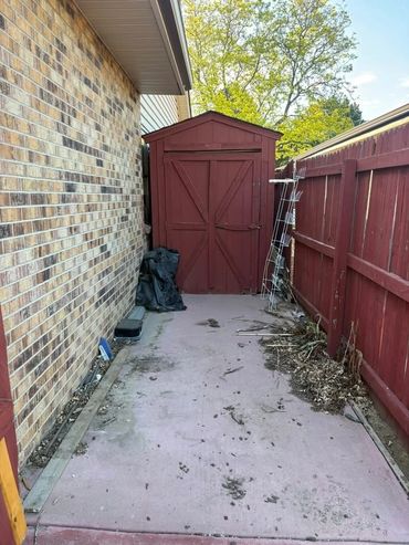 Outdoor red shed between a brick house and a red wooden fence with some debris on the ground.