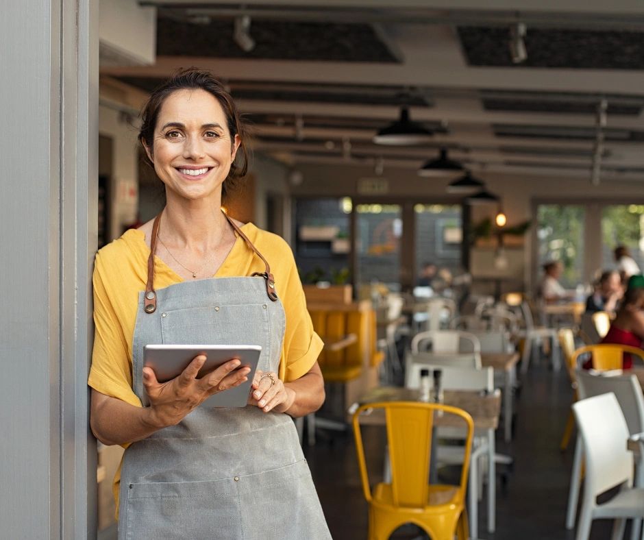 Smiling waitress in apron holding a tablet in a modern cafe.