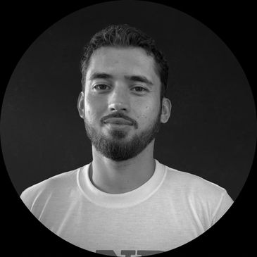 Young man with beard and white t-shirt smiling slightly against dark background.
