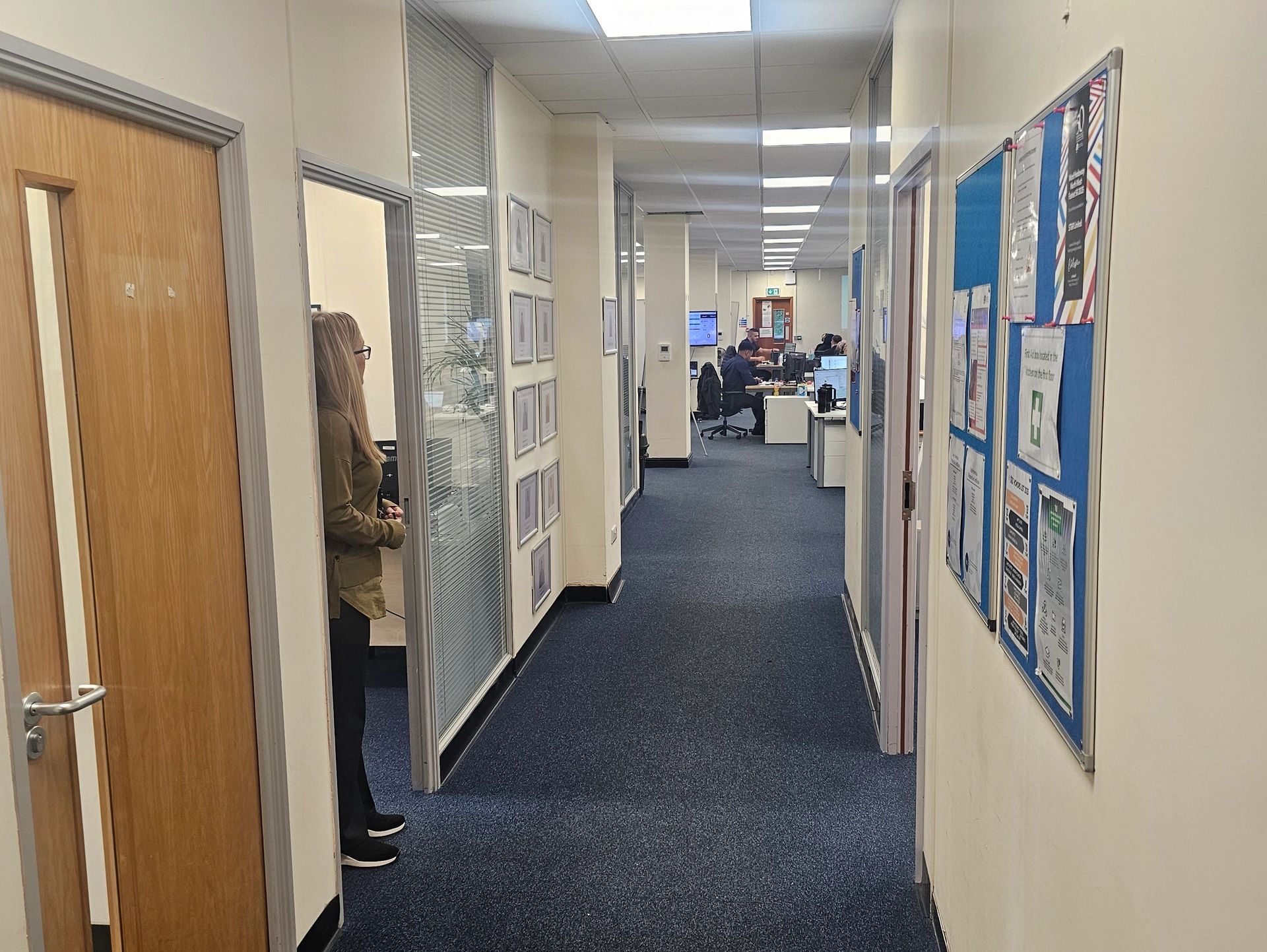 Office corridor with people working and a woman standing by a doorway.