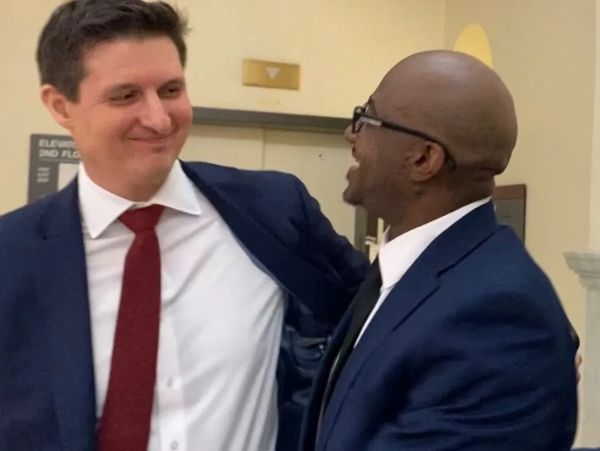 Two men in suits share a warm, joyful moment indoors.